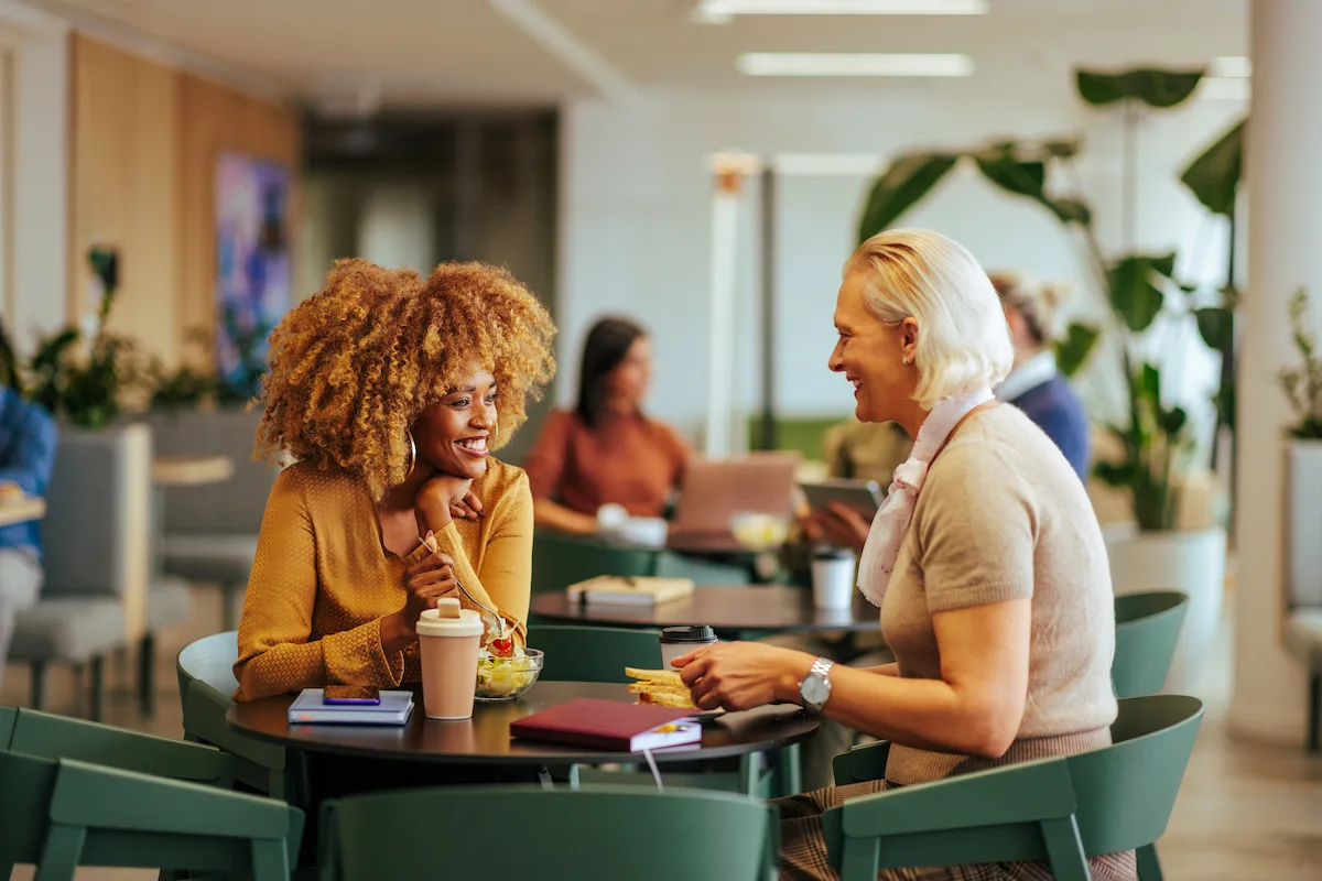 Two diverse coworkers are in the office lobby having breakfast and talking together.