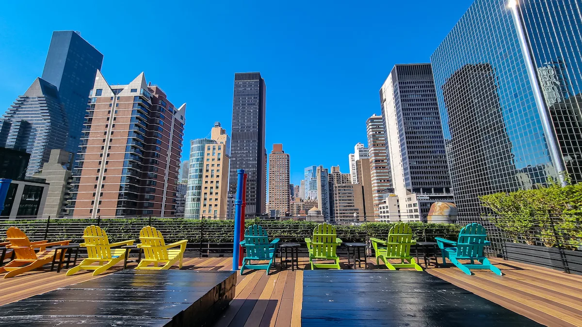 A rooftop with the city skyline view and modern skyscrapers against a sunny blue sky in New York. Colorful chairs overlooking the city. Rows of skyscrapers in the back. Sunny and warm day.