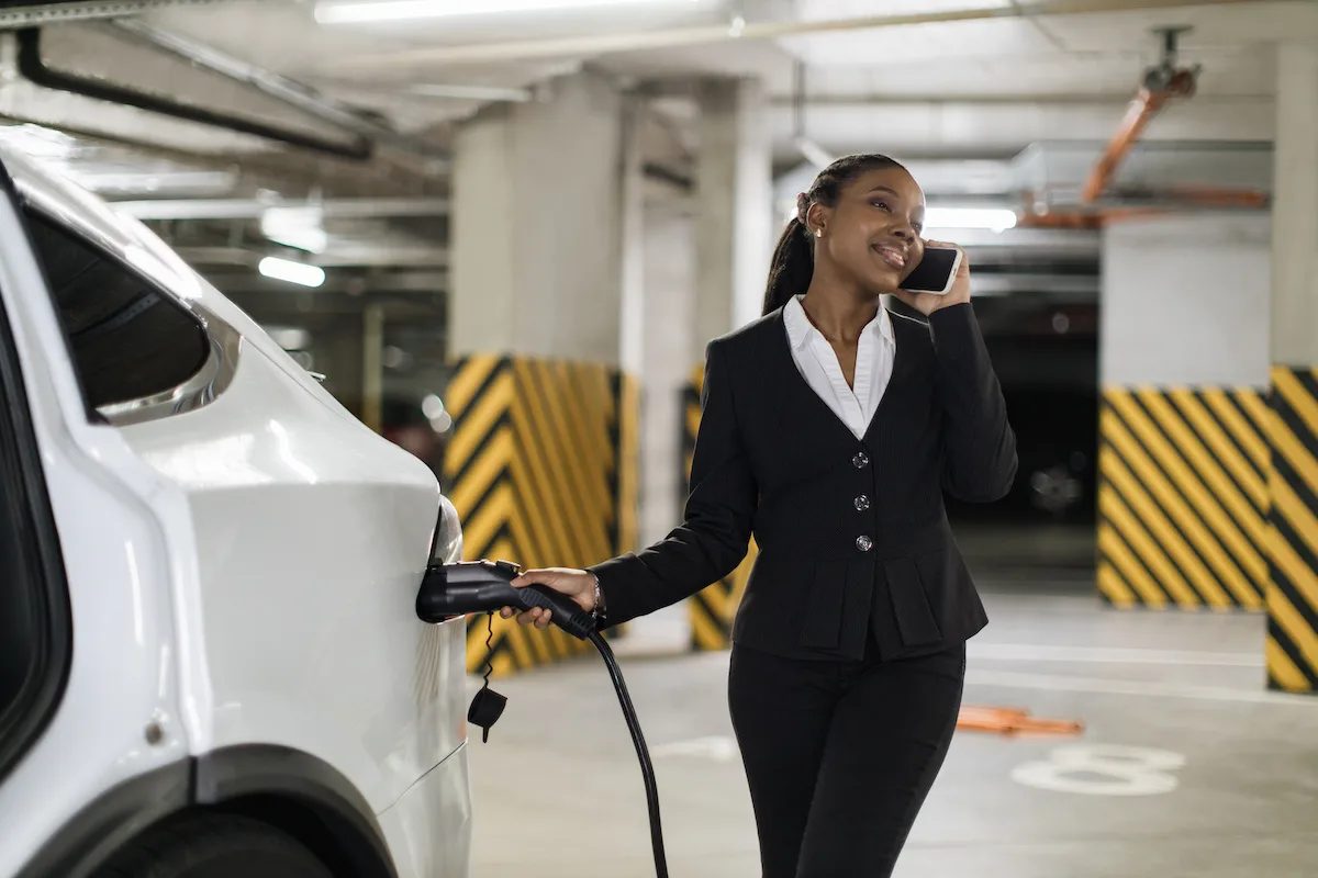 Cheerful woman having pleasant conversation on mobile while holding electric connector in e-car's port. Smiling african office worker providing contact details while dealing with charging process.
