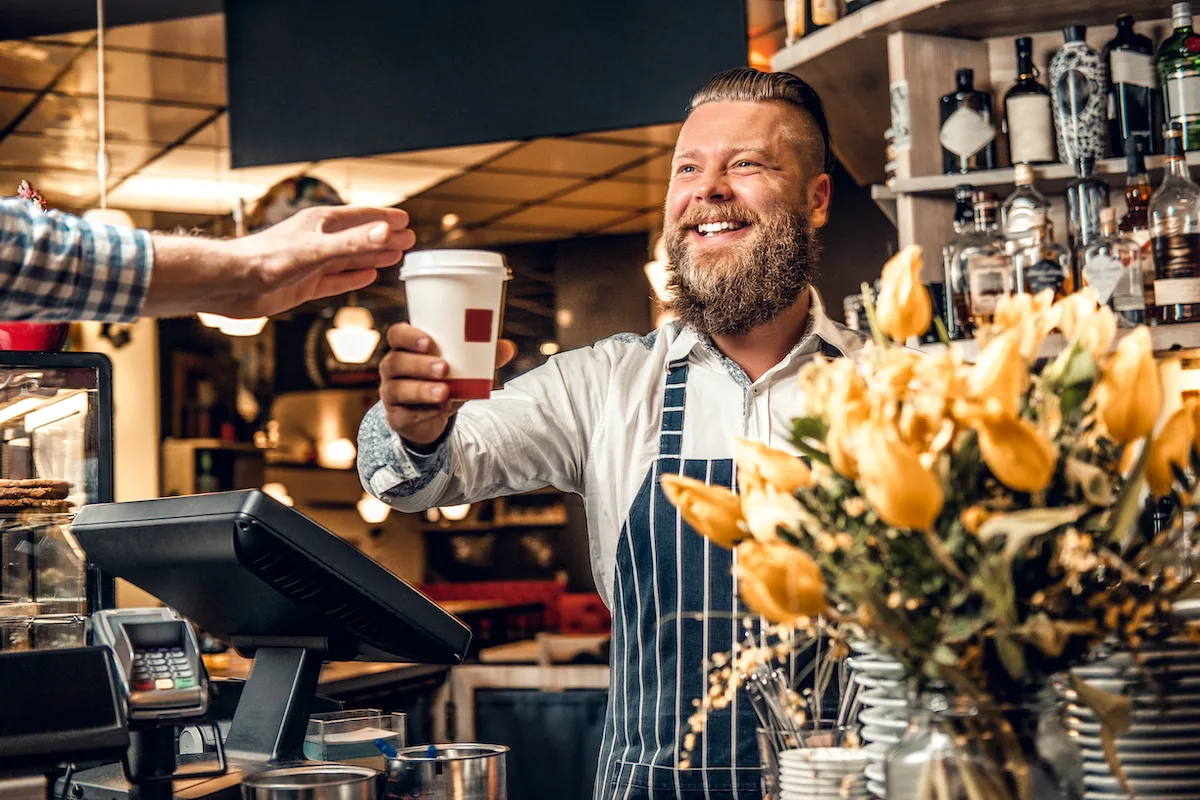 Positive bearded barista male selling coffee to a consumer in a coffee shop.