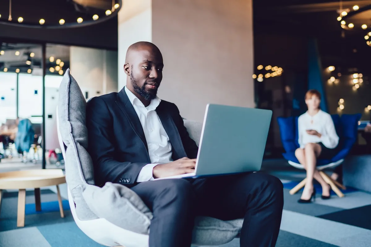 Busy African American male office worker in formal suit sitting on armchair and using laptop at stylish lobby of business center