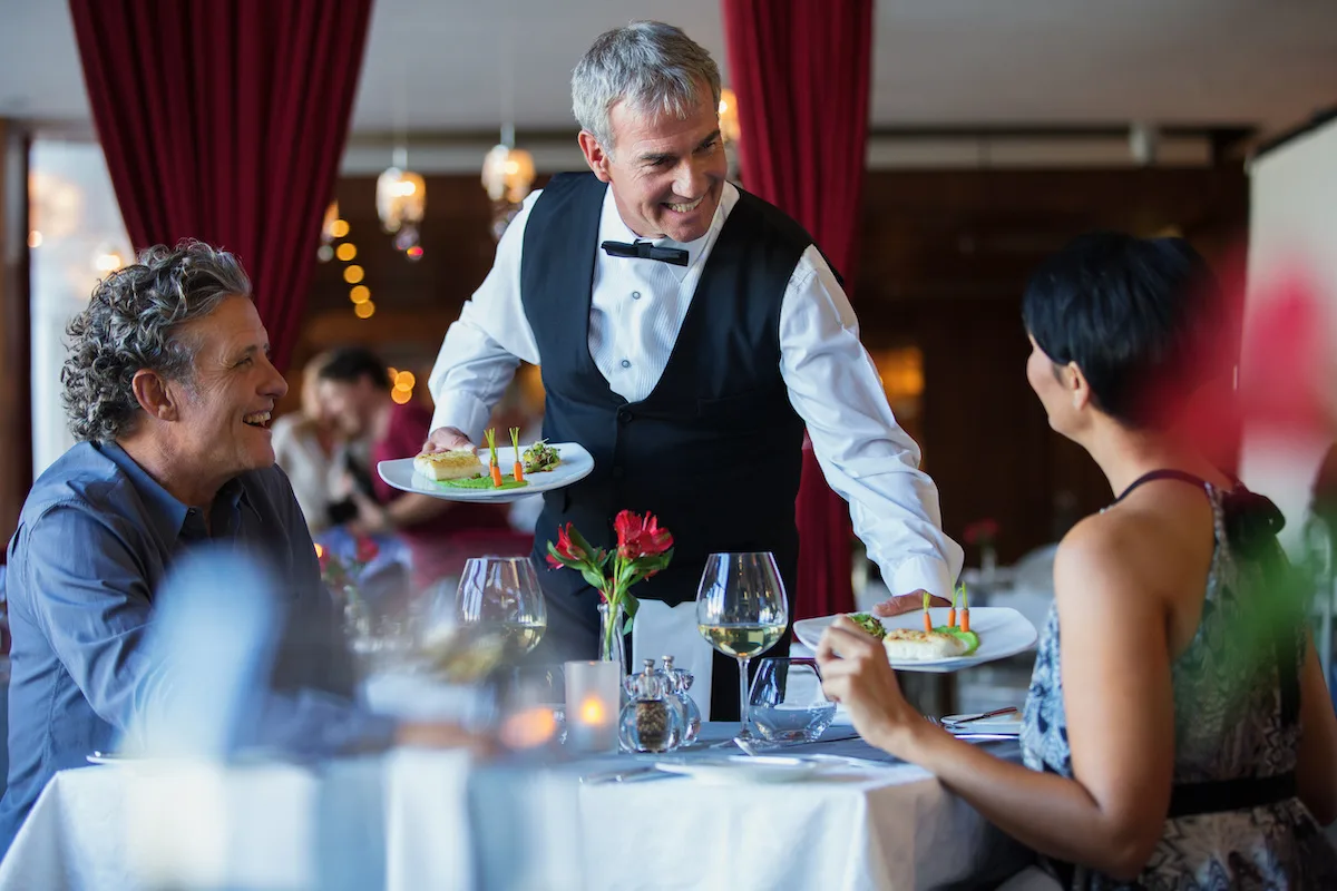 Smiling waiter serving fancy dishes to mature couple sitting at table in restaurant